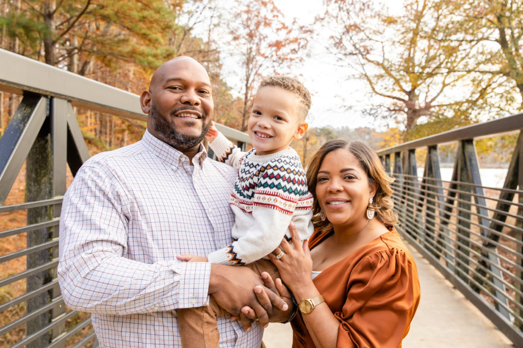 colored parents on white and brown outfits standing on a bridge with toddler boy in dad arms during extended family photo session in GA