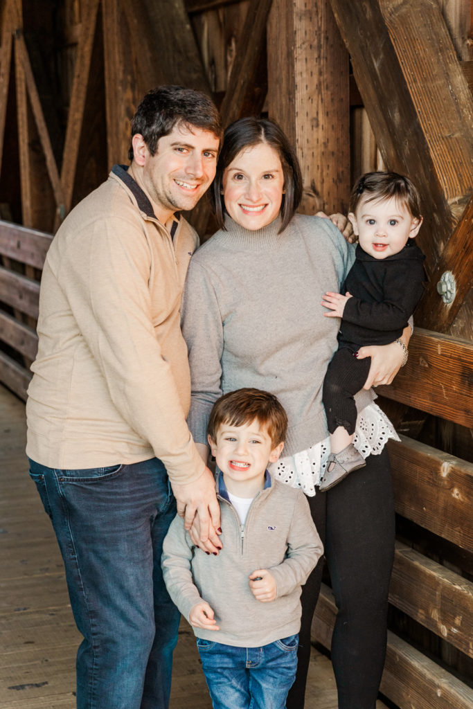 Milestone portrait session Alpharetta parents holding baby and toddler standing under a covered wooden bridge in fall