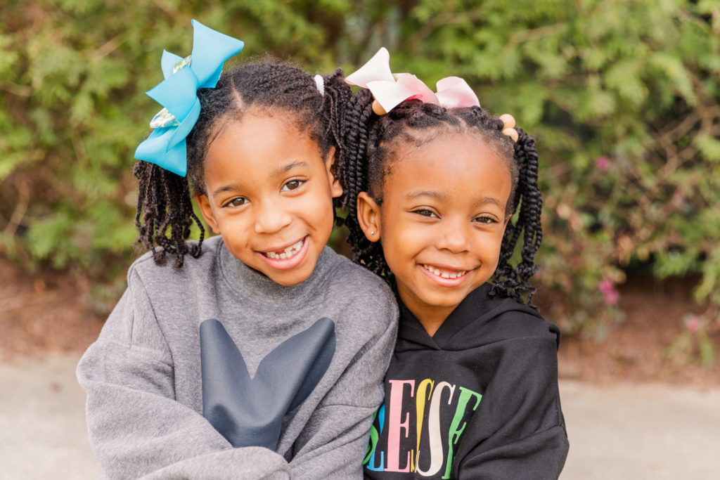 Toddler girls headshots with bows hugging and smiling sitting outside by Laure Photography in Atlanta