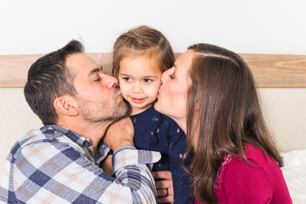 parents kissing toddle sitting on bed during indoor during lifestyle family photoshoot by Laure Photography