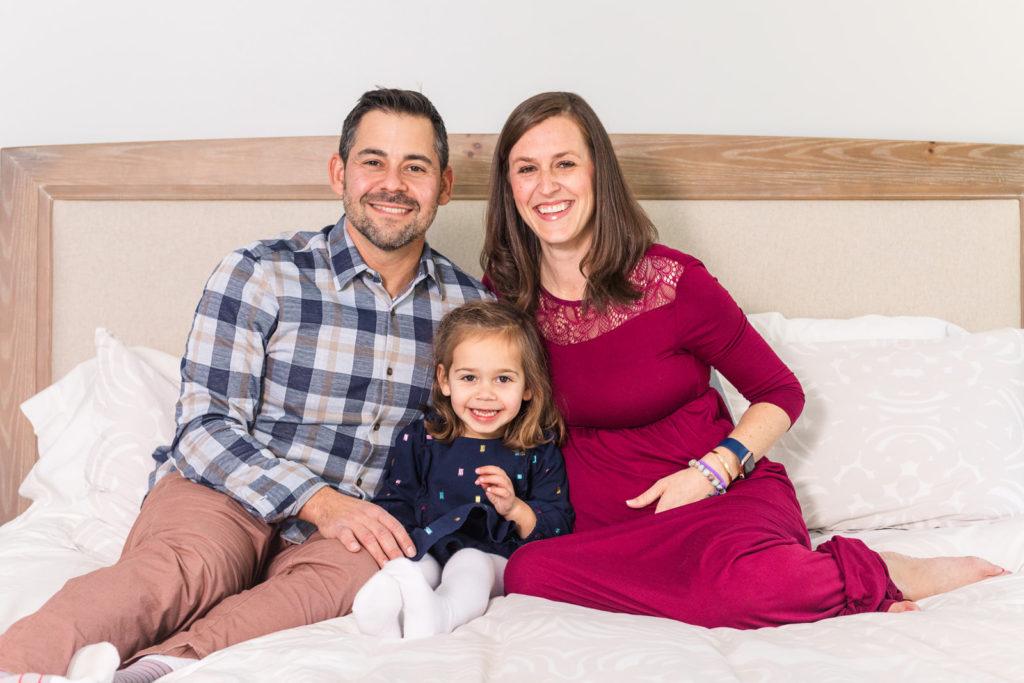 pregnant family with toddler girl sitting on a bed, daughter in blue dress and mum in maternity long pink dress with Atlanta photographer Laure Photography