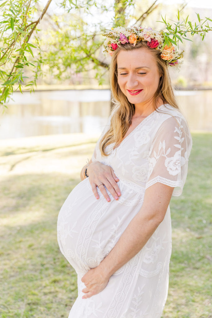 Pregnant woman looking down hands on her belly wearing a long white lace dress and a flower crown in Atlanta Duck Pond park with Laure Photography