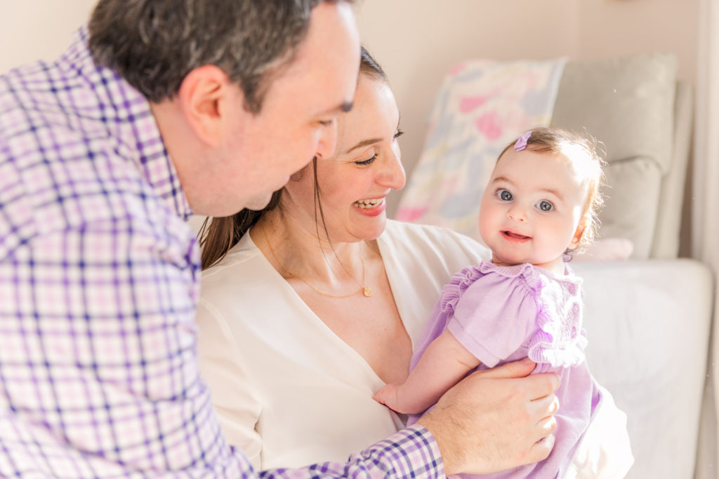 parents smiling and laughing with six month old baby girl wearing purple clothes baby looking at the camera with Atlanta Laure Photography