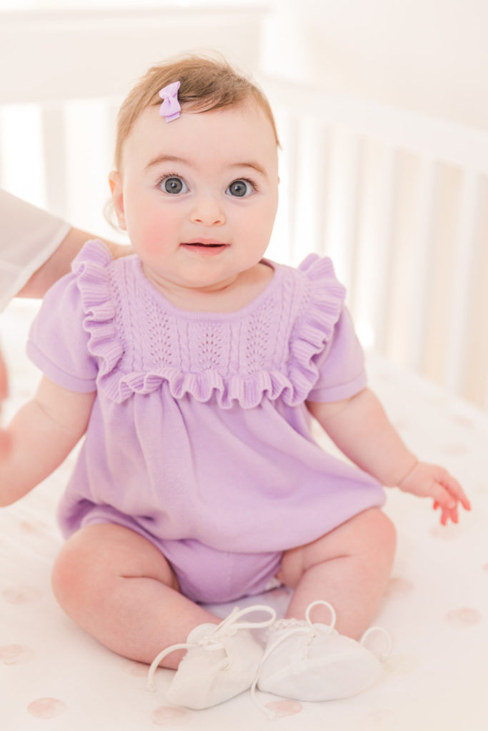 6 month old baby girl sitting on her bed in her nursery wearing a purple outfit with Atlanta Laure Photography