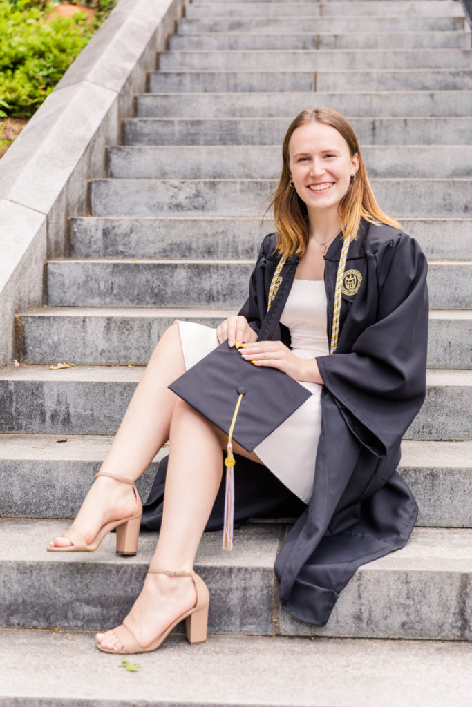 graduate woman in grown wearing a white dress and holding her cap sitting on steps on Georgia Tech Campus Atlanta by Atlanta photographer Laure Photography