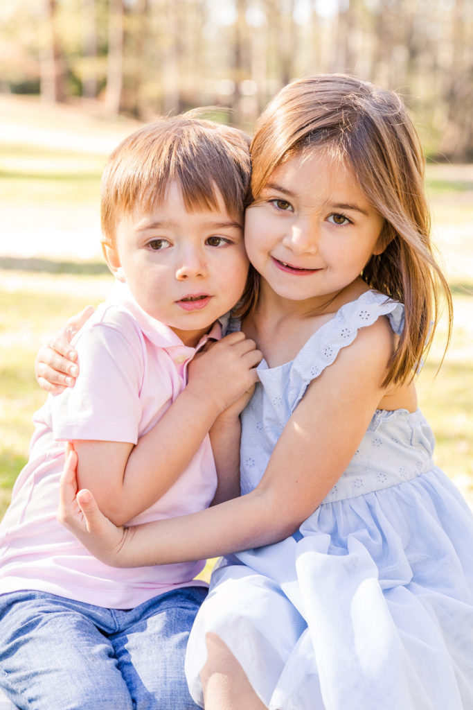 sibling boy in jeans and pink polo and girl in light blue dress sitting and hugging each other in a park with Laure Photography Atlanta