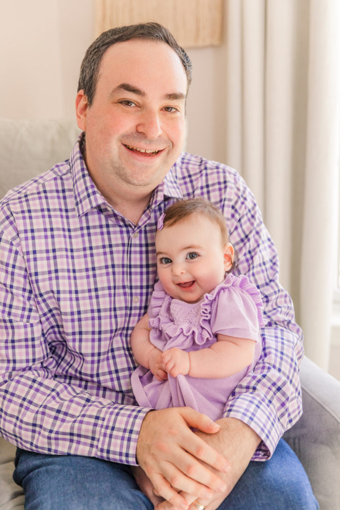 daddy sitting with six month old baby girl on his laps both looking and laughing in coordinated purple and white outfits with Atlanta Laure Photography