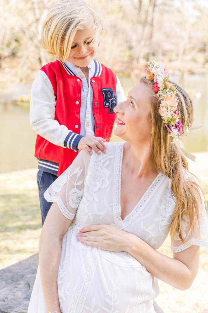 pregnant woman sitting hands on her belly looking at her son who is standing behind her she is wearing a long white lace dress and a flower crown in Atlanta Duck Pond park with Laure Photography