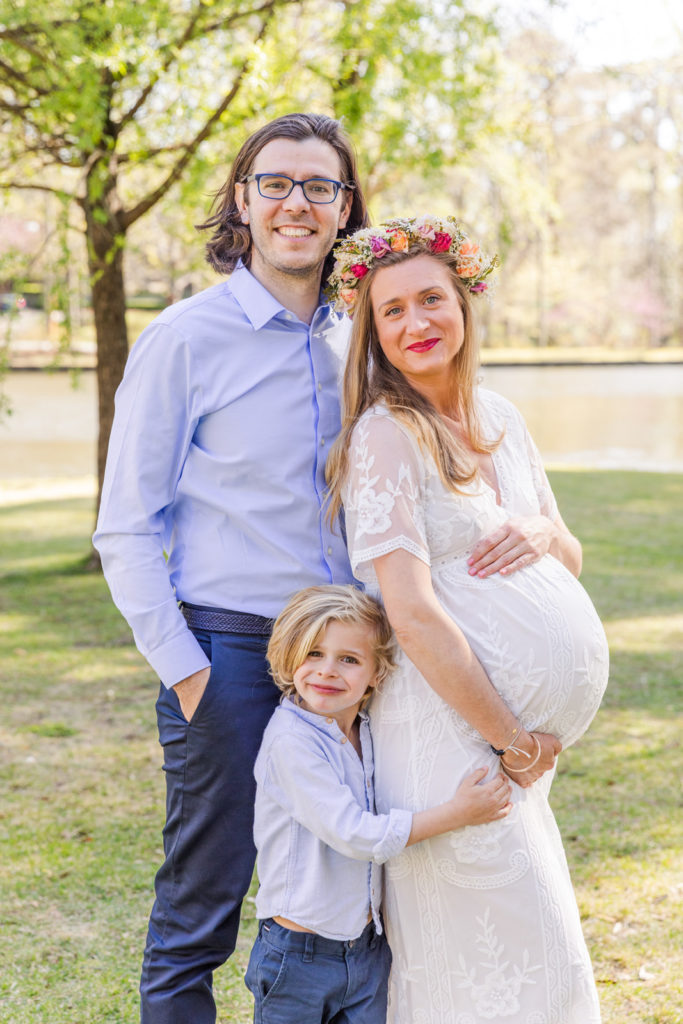 pregnant family with dad mom and son standing dad and son wearing corrdinated blue pants and shirts and mom wearing long white lace dress and a flower crown in Atlanta Duck Pond Park by family photographer Laure Photography