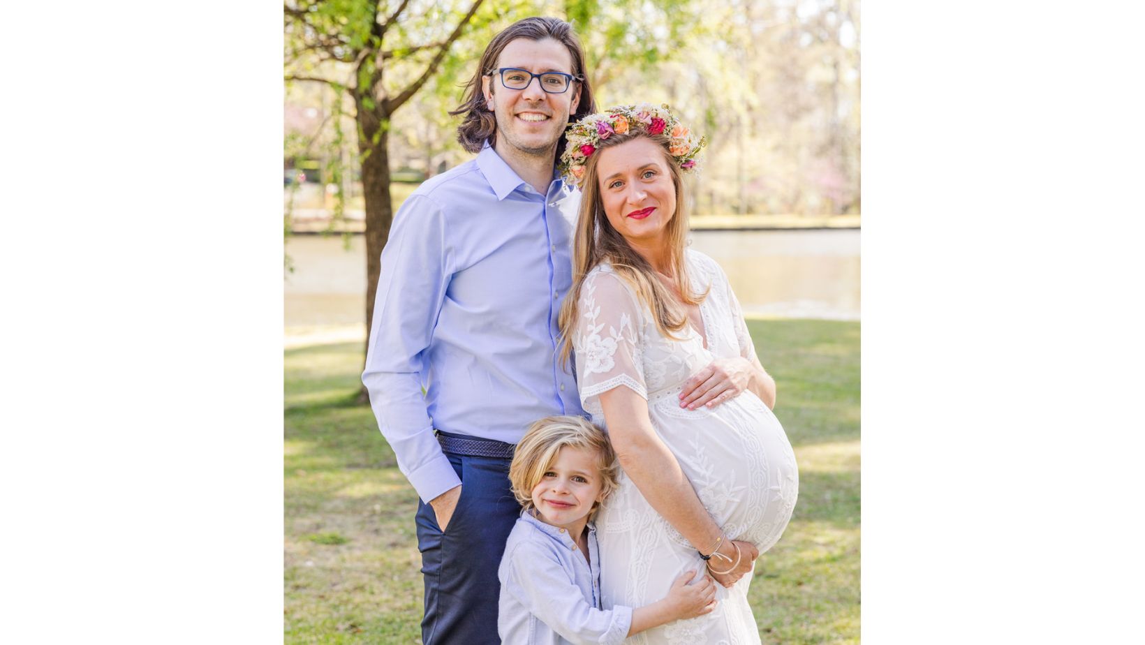 pregnant family with dad hands in pockets mom hands on pregnant belly and son standing dad and son wearing corrdinated blue pants and shirts and mom wearing long white lace dress and a flower crown in Atlanta Duck Pond Park by family photographer Laure Photography