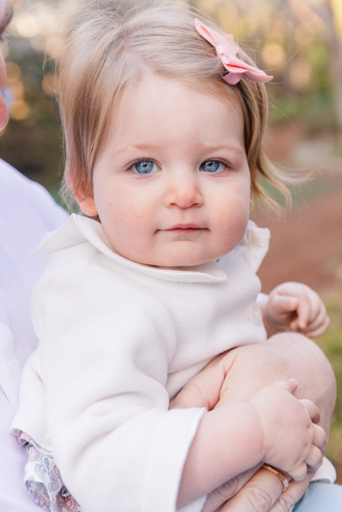 baby one year old girl with blue eyes wearing a white jacket and a pink bow baby girl sitting on baby wooden chair on front of a ONE wooden sign on the floor in front a pond on a park with Laure photography Atlanta