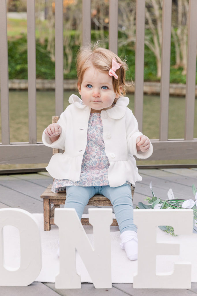 baby girl with white jacket and flower printed top and pink bow sitting on baby wooden chair on front of a ONE wooden sign on the floor in front a pond on a park with Laure photography