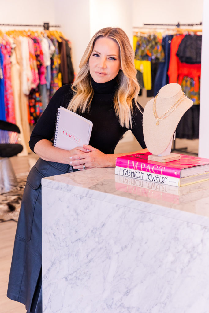 woman wearing black turtle neck and black leather skirt leaning agains a fashion store counter holding a notebook