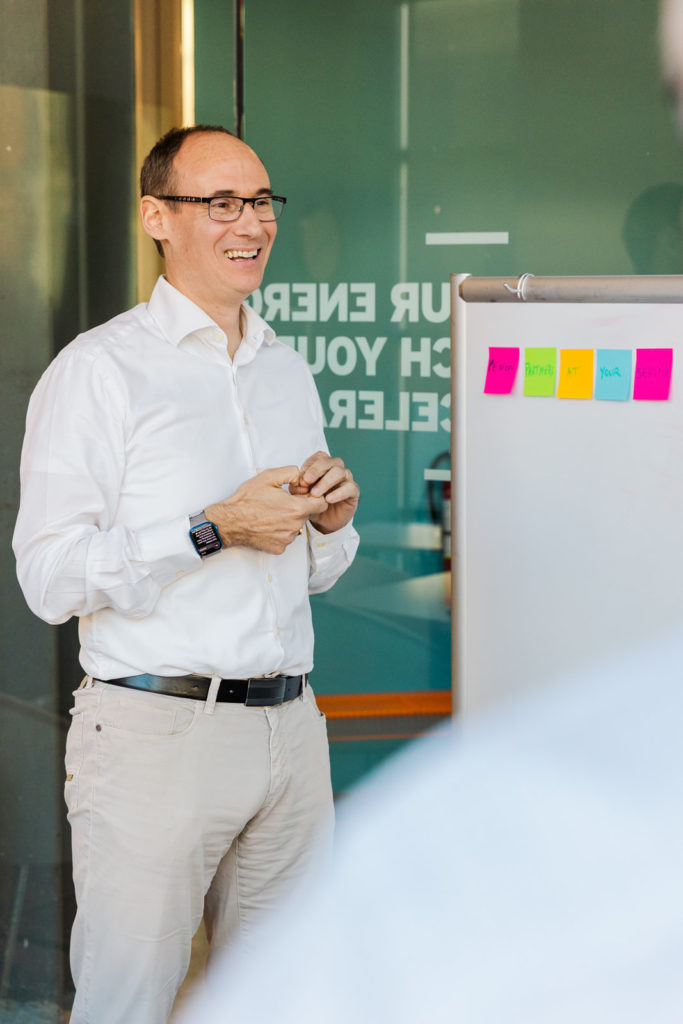 man in white shirt and beige pant standing in front of a white board with post its talking to someone during a team meeting in a conference room Laure Photography brand shoot