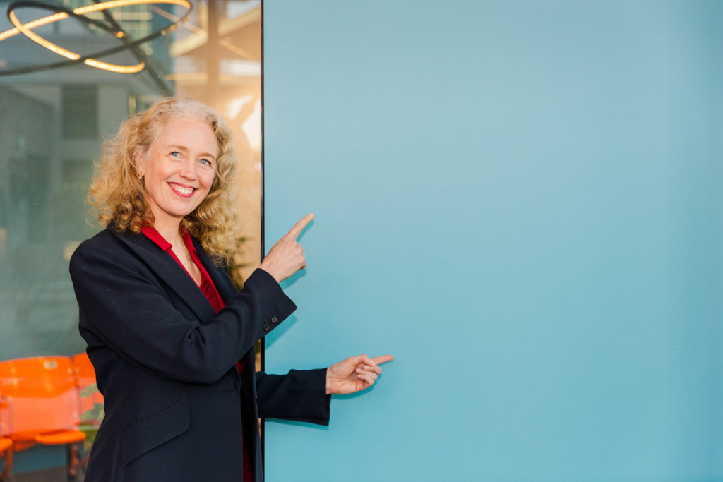 Woman wearing red top and black jacket pointing at a blue negative space board with both hands during brand shoot for website announcement Laure photography Atlanta