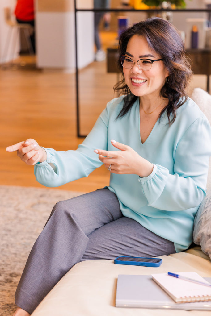 woman wearing light blue silk top and gray pant sitting legs crossed on a sofa talking and smiling to a person off camera brand photo session Laure Photography