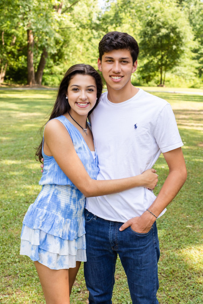 Teenager siblings brother and sister hugging in a park teenager girl wearing a white and blue dress and teenage boy wearing white Ralph Lauren tee-shirt and blue jeans standing in a Sandy Spring park in GA Laure photography