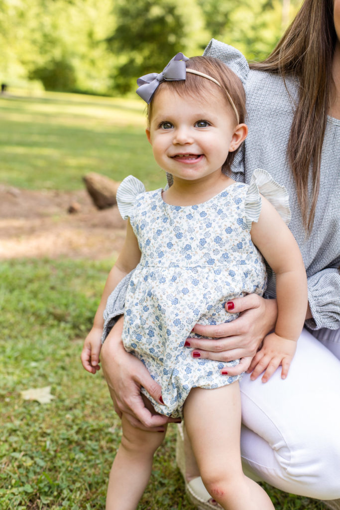 toddler girl wearing a white printed jumper with blue flowers and a coordinated blue bow smiling leaning agains her mom during family photo session Laure Photography