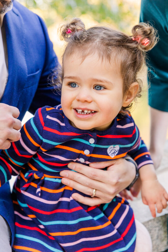 toddler girl smiling off camera wearing a navy blue dress with orange pink and light blue stripes