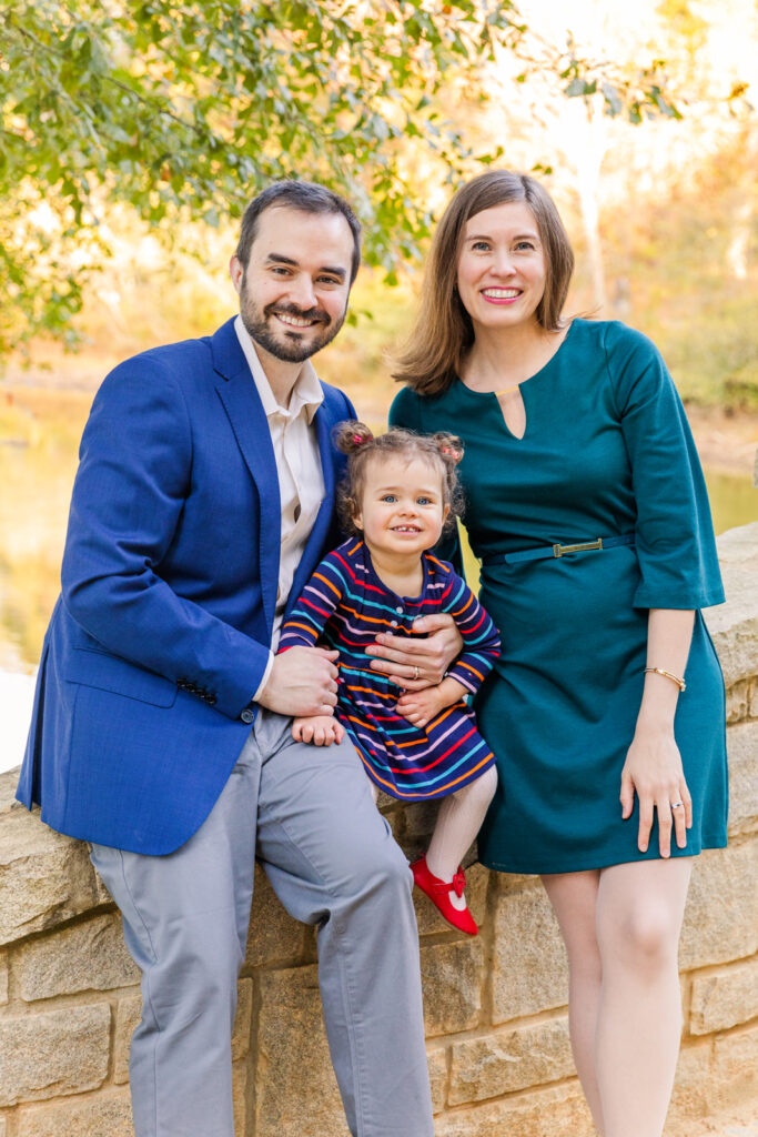 family with toddler girl sitting on a wall in front of a lake in an Atlanta park wearing coordinated blue and green outfits
