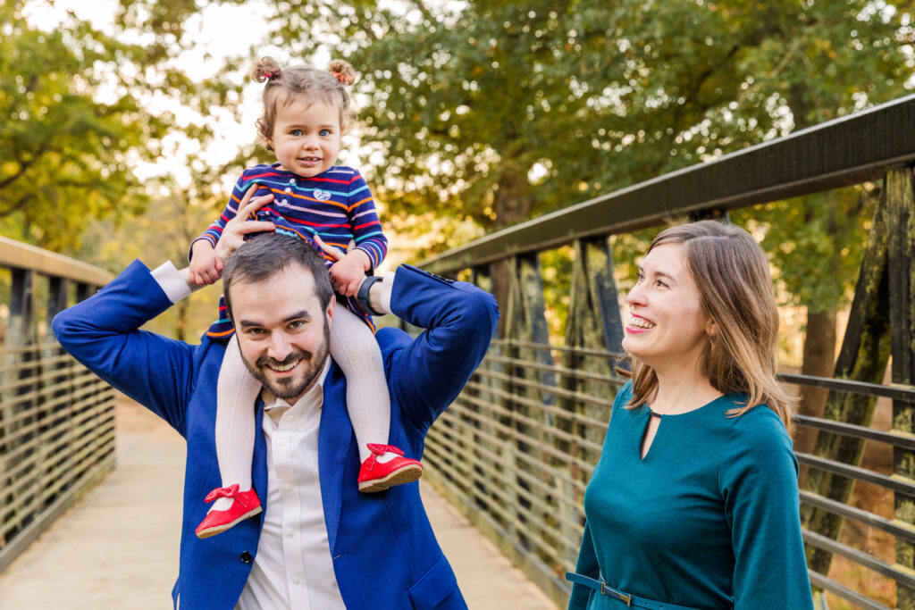 lifestyle photo of ad holding toddler girl on his shoulder and mom looking at them in a bridge in Atlanta