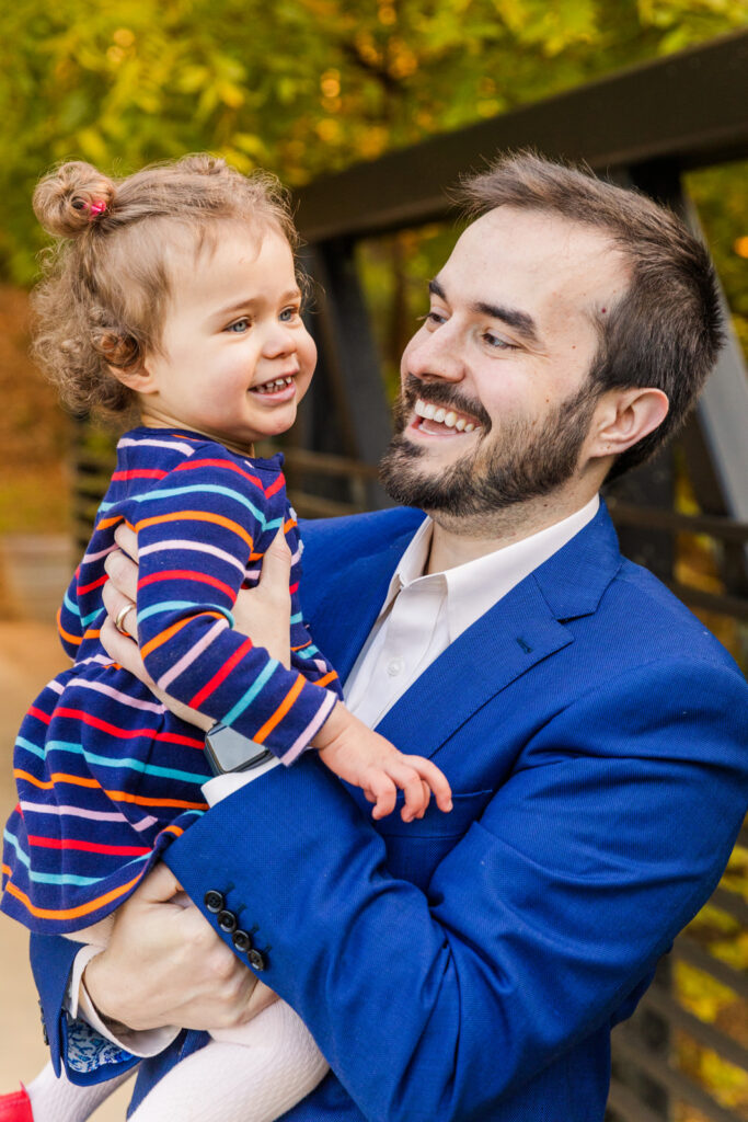 dad holding toddler girl in his arms and laughing in a park wearing coordinated blue outfits