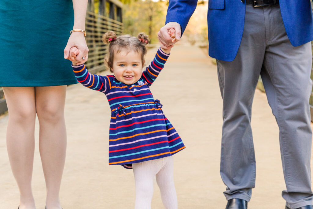 toddler girl standing and holding her parents hands in a park family wearing coordinated blue and green outfits