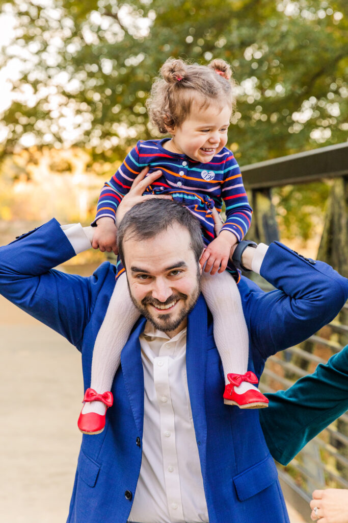 Dad holding toddler girl on his shoulders with daughter laughing off camera wearing coordinated outfits in an Atlanta park