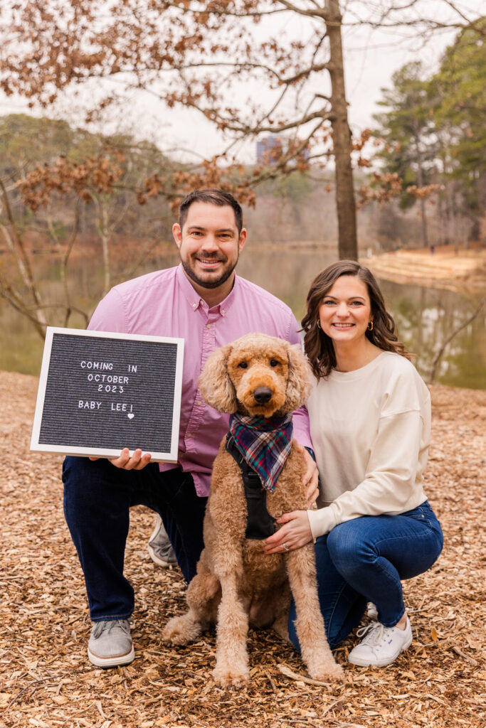 Couple with their dog holding a baby announcement sign in front of a pond by Atlanta family photographer Laure Photography