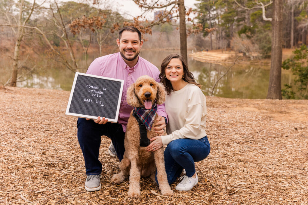 Couple with their golden-doodle dog holding a baby announcement sign in front of a park pond by Atlanta family photographer Laure Photography
