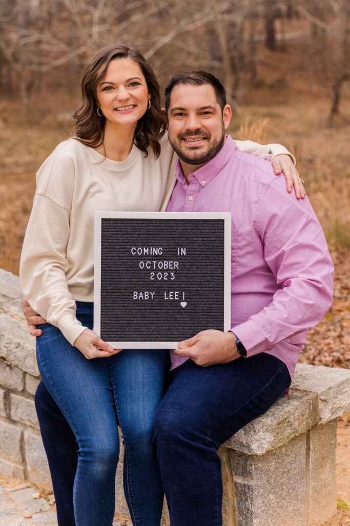 Couple sitting and holding a baby announcement sign in a park by Atlanta family photographer Laure Photography