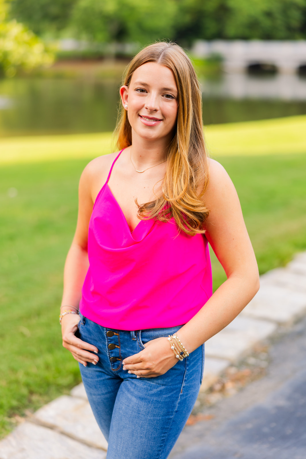 teenager wearing blue jeans and a pink top standing ankles crosses and hands in pockets in front of a pond and a bridge in Buckhead park during senior portraits