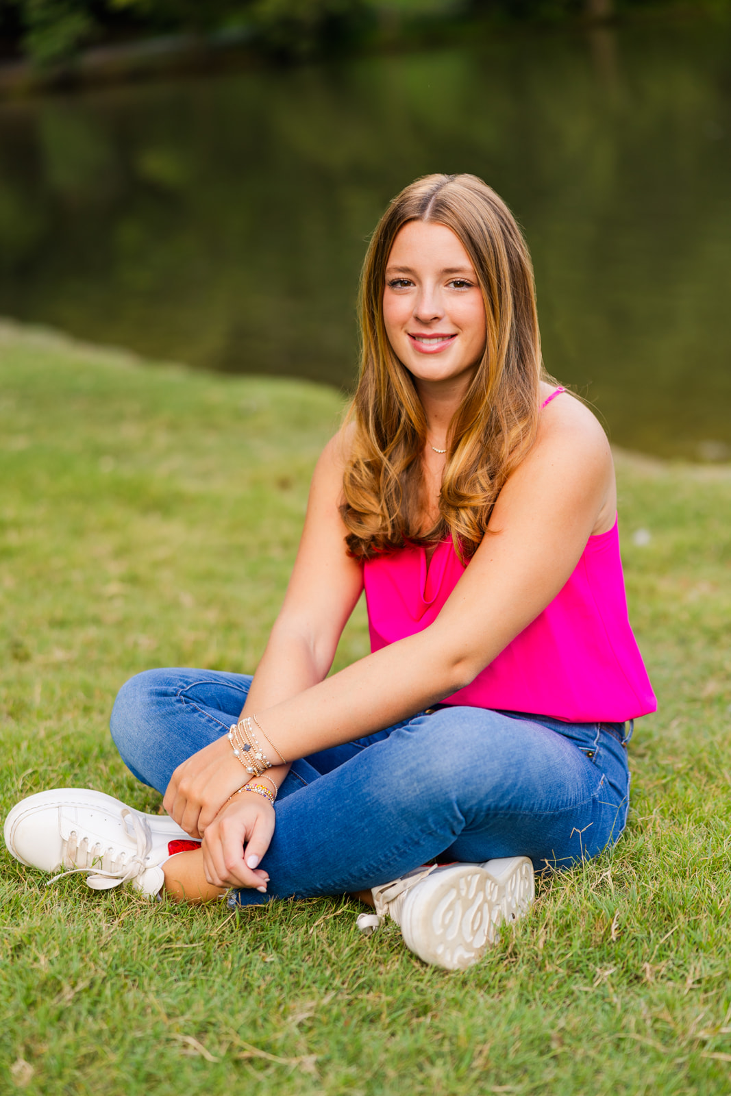 teenager sitting criss cross on the grass wrists crossed in front of a pond in Buckhead during senior portraits