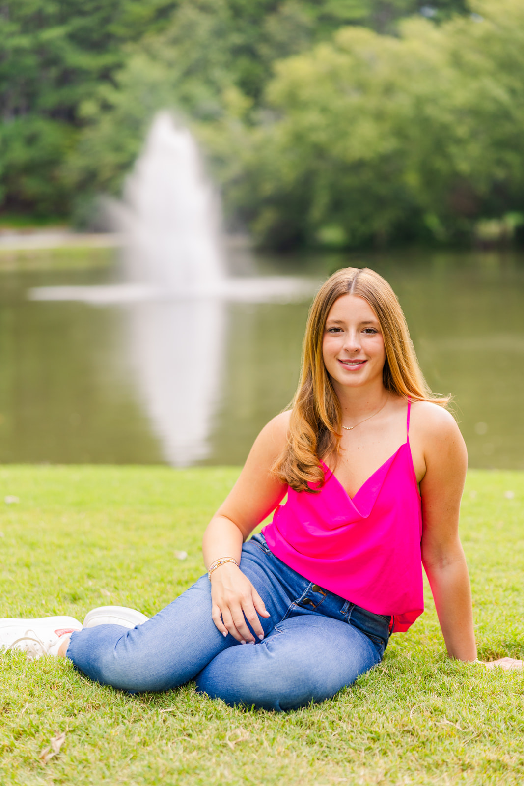 young adult wearing jeans sitting legs on the side on the grass in front of a fountain during senior portraits in Atlanta GA