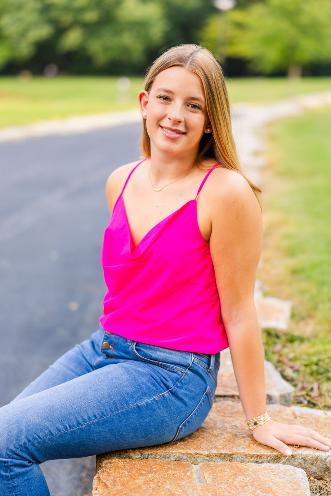 high school girl wearing jeans pants and a pink top sitting on the side of a path during senior photo session in an Atlanta GA park