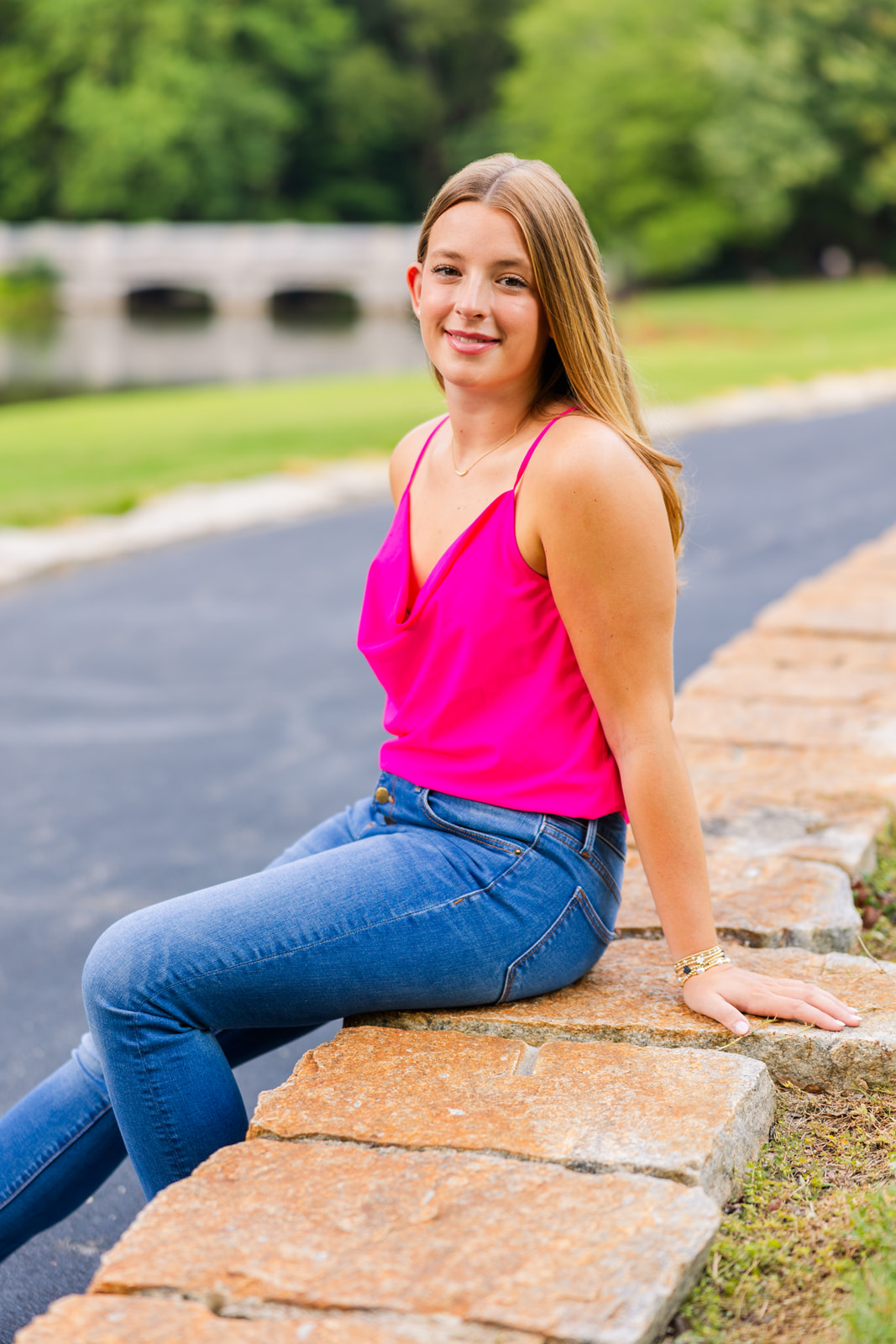 high school girl wearing jeans and a pink top sitting in front of a bridge during senior photoshoot in Buckhead GA