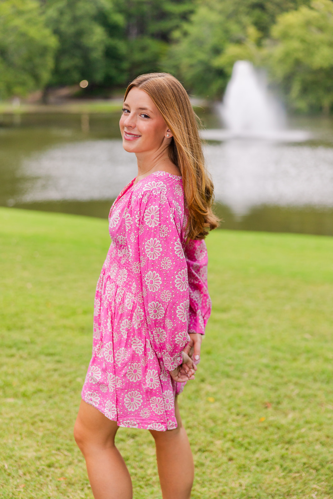 senior girl in a pink and white summer dress with standing sideway in front of a fountain in Lenox park during portraits