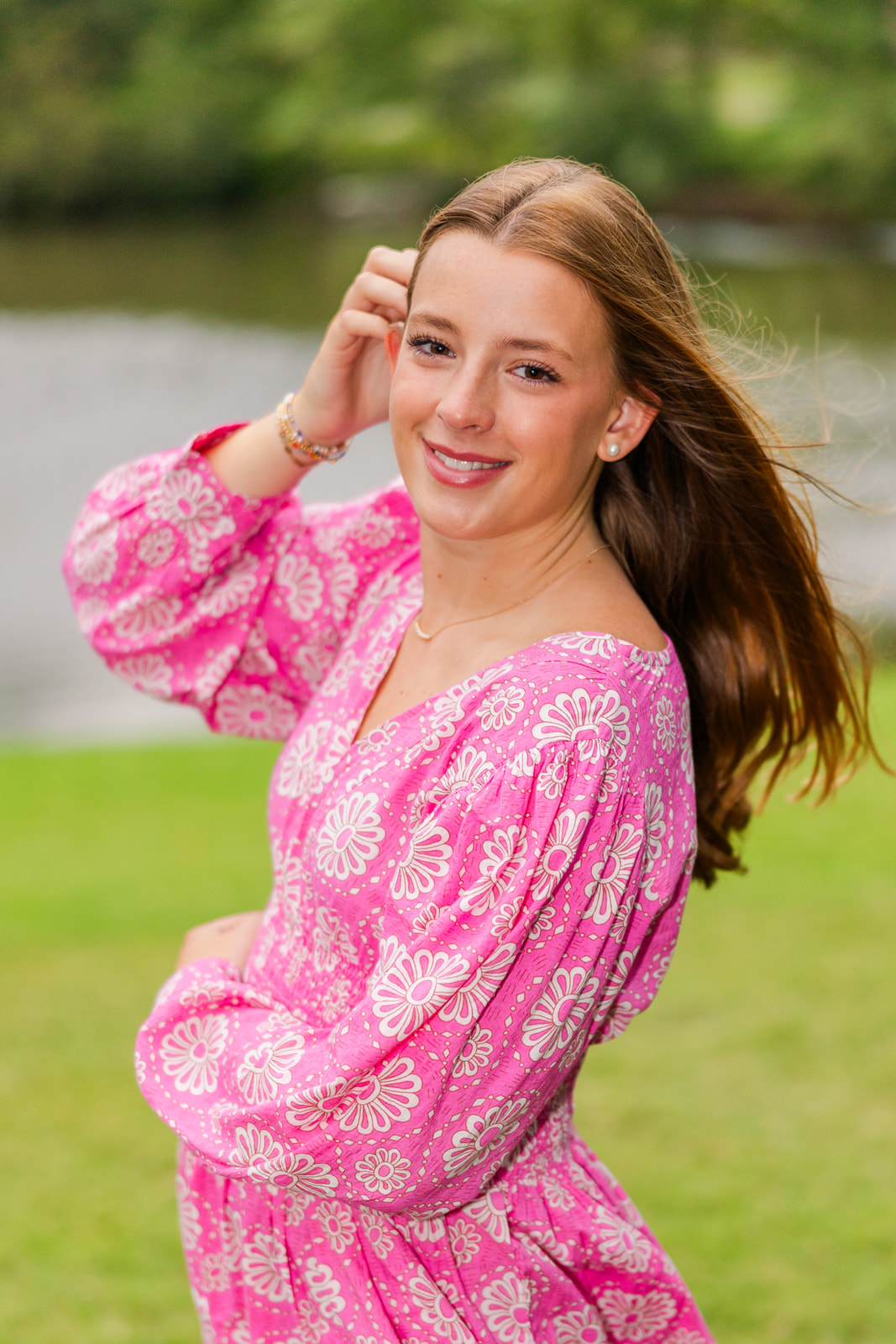 teenage girl in a pink dress with white flowers patterns standing sideway a hand in the hair in front of a pond in Atlanta park during photos