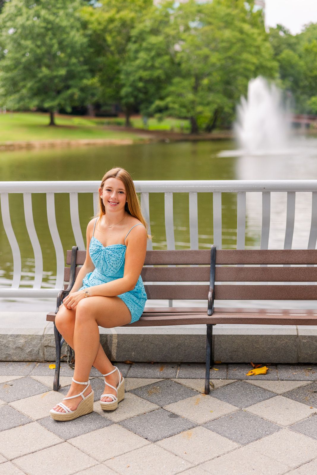 senior girl sitting sideway on a bench on a bridge in front of a fountain weary a light blue romper during her portraits photoshoot in Atlanta Lenox Park