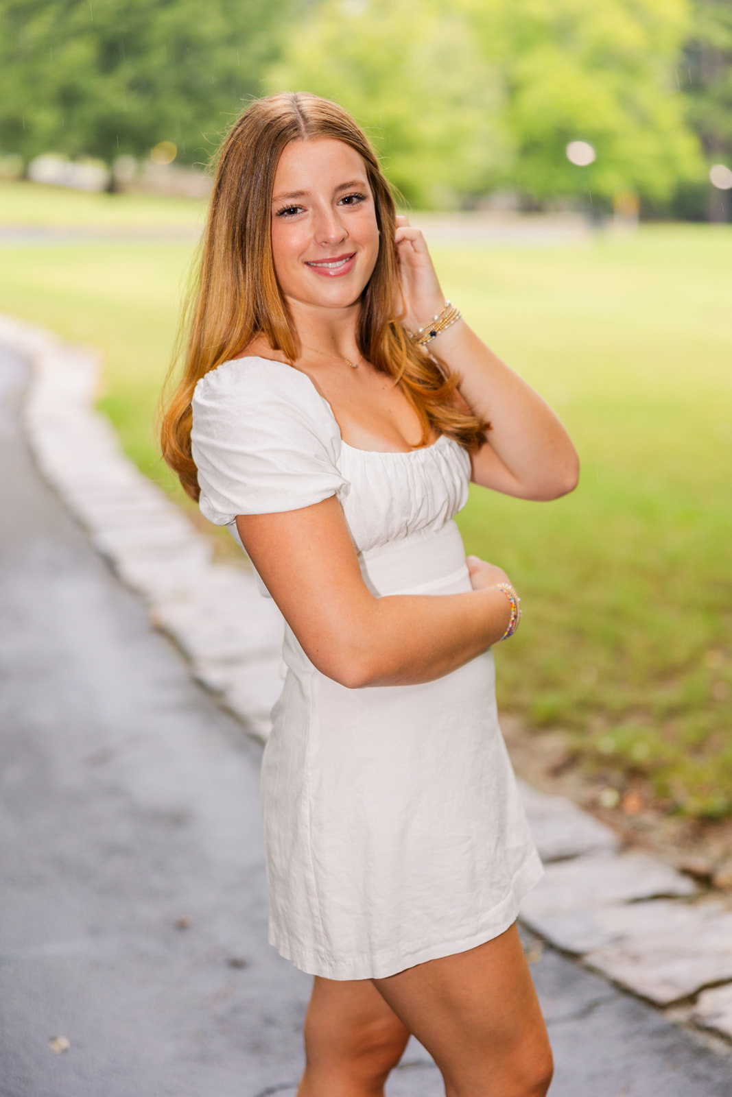 teenage girl standing side way one hand in her hair wearing a white short dress in an Atlanta Park during Senior portrait photo session