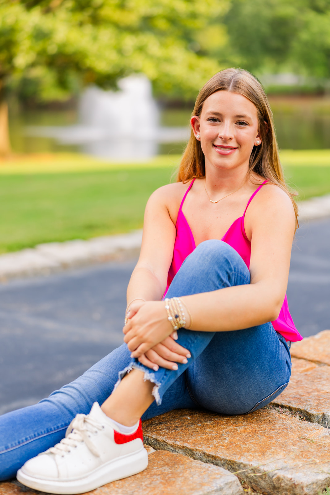 young adult sitting in front of a fountain in an Atlanta park hugging a knee wearing jeans and pink sleeveless top during senior photoshoot