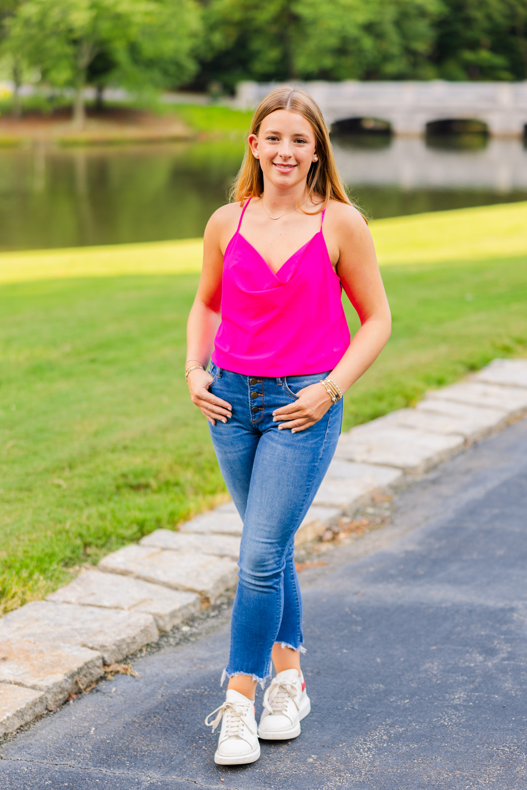 teenager wearing jeans, sneakers and pink top standing ankles crosses hands in pockets in front of a pond and a bridge in Buckhead park during senior photos