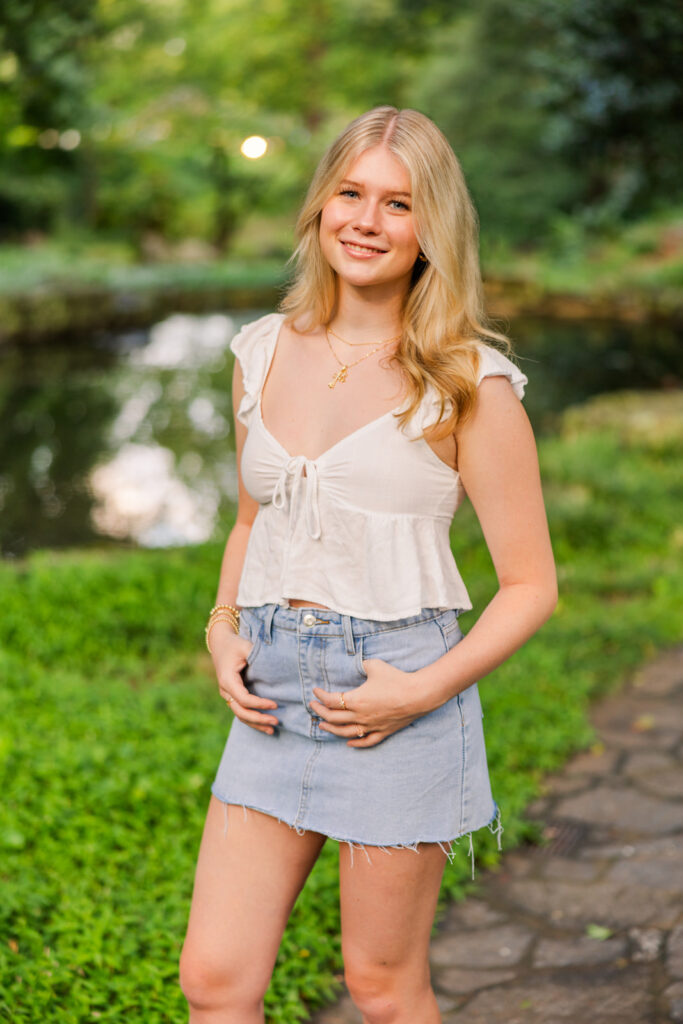 senior girl standing ands in pocket of jeans skirt smiling at the camera in front of Winn park pond in Midtown Atlanta GA
