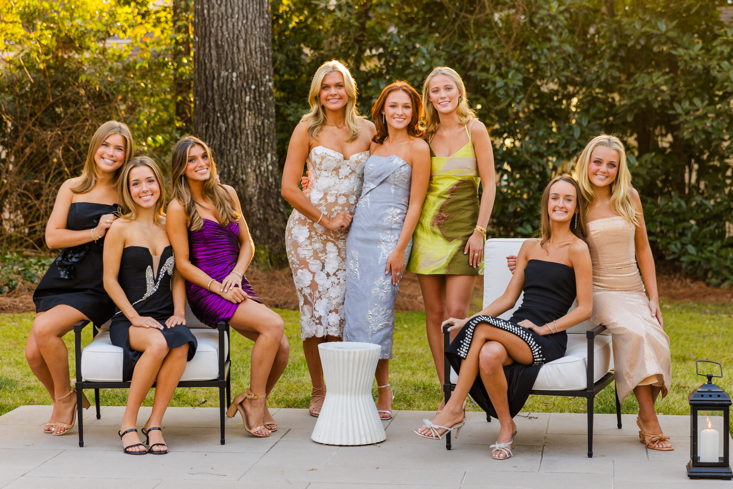 group of high school Senior girls from high school in party long dresses posing sitting and standing in front of a greenery background