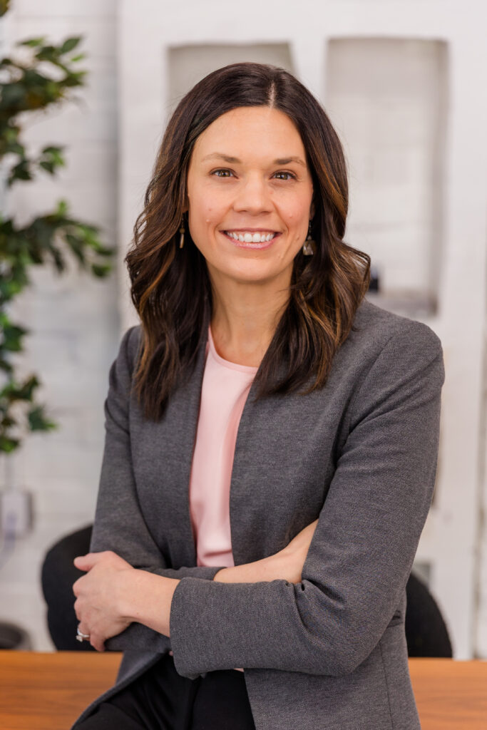 woman with long brown hear leaning against a desk arms crossed wearing corporate outfit light pink top and gray jacket during headshot photoshoot with Laure Photography