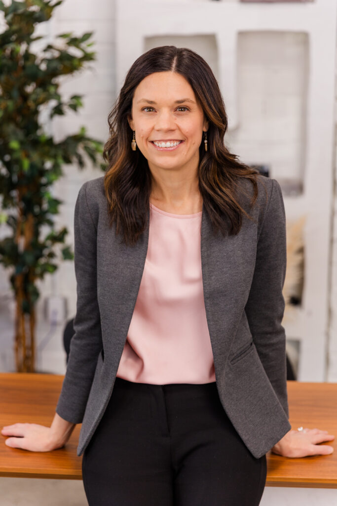woman leaning against a desk one hand on each side wearing corporate outfit light pink top and gray jacket during headshot photoshoot with Laure Photography