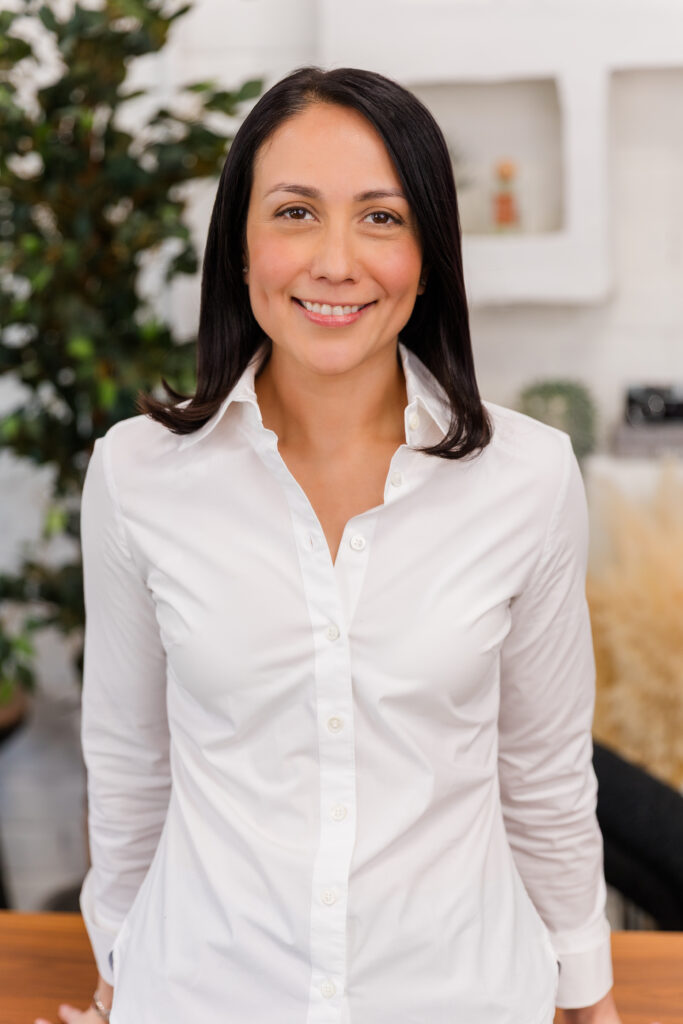 woman standing in front of lifestyle background with office and greenery props leaning against a desk one arm on each side wearing corporate outfit white tee-shirt and dark blue jacket during headshot photoshoot with Laure Photography