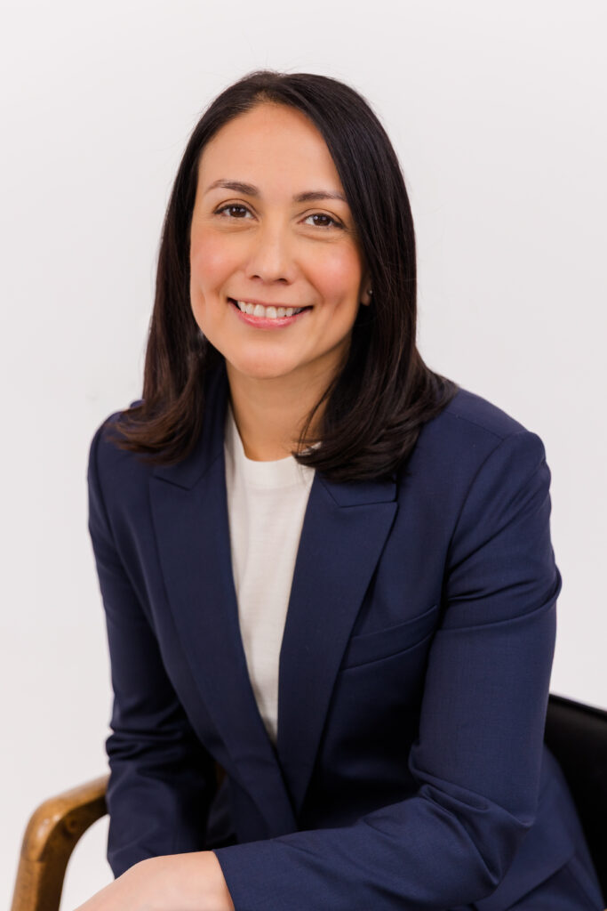 woman sitting on a chair in front of white background wearing corporate outfit white tee-shirt and dark blue jacket during headshot photoshoot with Laure Photography