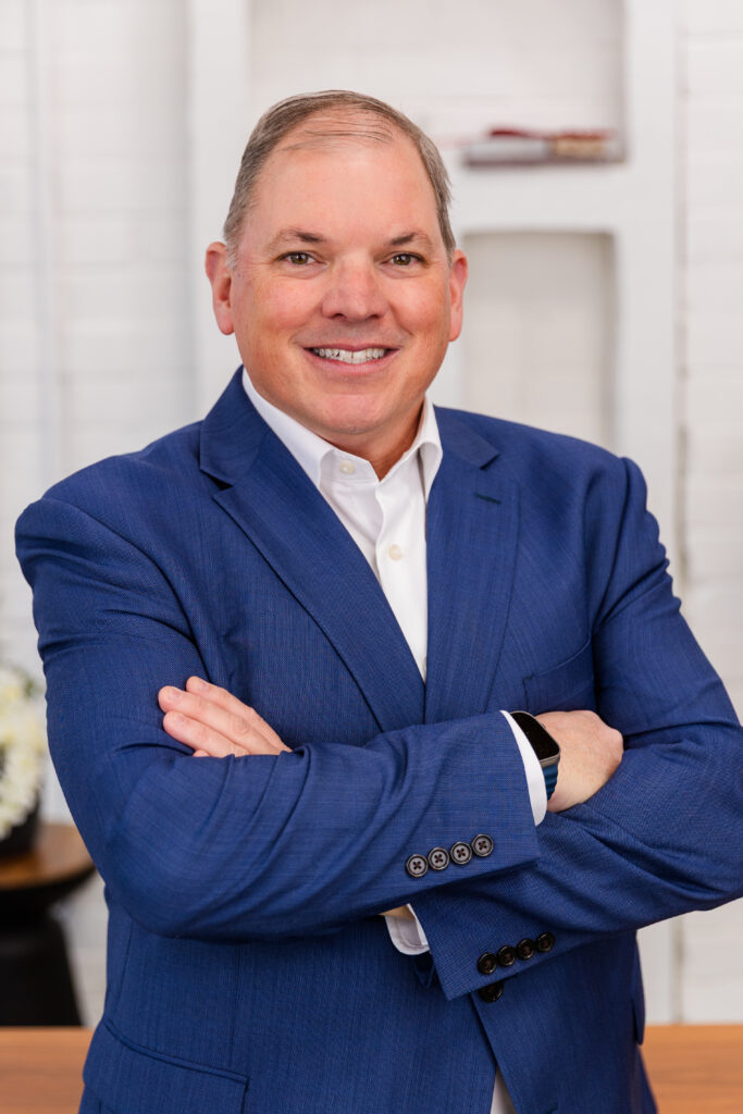 man standing arms crossed in front of a desk with a lifestyle office background wearing corporate outfit white shirt and blue jacket during headshot photoshoot with Laure Photography