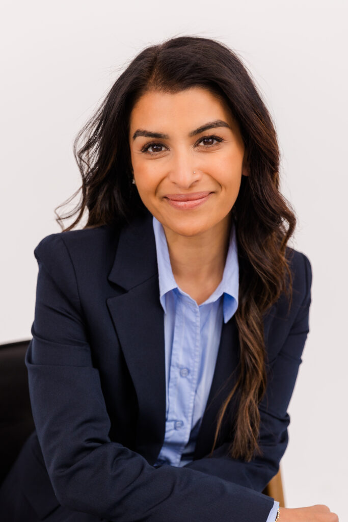 woman with long brown hair sitting on a chair in front of white background wearing corporate outfit light blue shirt and dark blue jacket during headshot photoshoot with Laure Photography